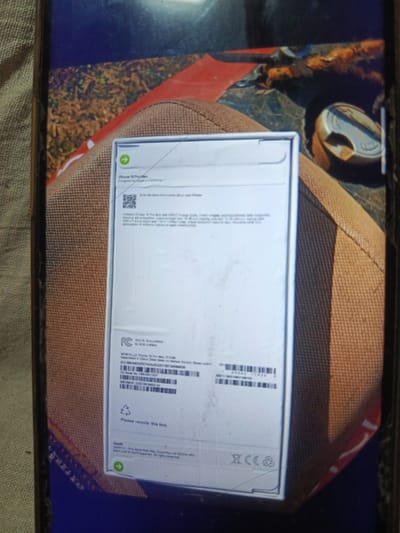 The image shows a close-up of an iPhone 15 Pro Max box. The box is white with text and a QR code. The text indicates the product is an "iPhone 15 Pro Max" and it was "Designed by Apple in California". There are also regulatory markings, including "FC", "CE", and a recycling symbol. The box appears to be resting on a textured surface, possibly fabric, and is partially obscured by a red and brown object. The background is dark and out of focus, showing what appears to be natural elements like dirt and foliage. There is no information in the image that relates to the city of Svoboda, Russia.
