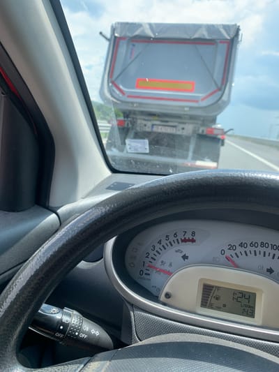 The image shows a view from the driver's seat of a car on a highway. In front of the car is a large truck. The dashboard of the car is visible, with a speedometer and a fuel gauge. The fuel gauge indicates the fuel level is low. The digital display shows a time of 1:24 and a fuel consumption of 14.3 L/100km. The scene appears to be on a sunny day with some clouds in the sky. The location is identified as the city of Rajana, Bulgaria.
