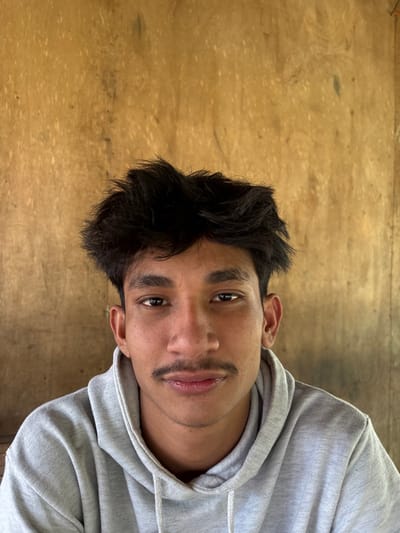 A young man with dark hair and a mustache is angled slightly toward the camera, wearing a gray hooded sweatshirt. His eyes are dark and his expression neutral. The background features a wooden surface. The image is well-lit, highlighting the texture of the wood and the details of the man's face and clothing.