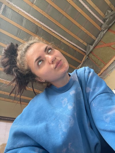 The image shows a close-up, low-angle shot of a young woman looking upwards and to the right. She has her hair pulled up in a messy bun and is wearing a blue tie-dye sweatshirt. The background appears to be the underside of a roof under construction, with wooden beams and insulation visible. There is no indication of the specific location within Мосметаллоконструкция Онт, Russia.