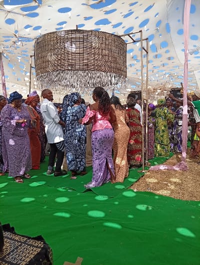 An outdoor social gathering is depicted in Kerang, Nigeria, beneath a white fabric canopy perforated with numerous holes through which the blue sky is visible. The ground is partially covered by a vibrant green carpet, which transitions to an area of dry straw or hay on the right. A large, cylindrical decorative light fixture, composed of dark woven material with silver fringed tassels, hangs centrally from a metal support structure. Approximately 15 to 20 adults are present, primarily dressed in colorful, patterned traditional attire, including numerous women wearing headwraps. In the foreground, a woman wearing an off-the-shoulder pink top and a form-fitting purple patterned skirt with a side slit is observed with her back partially to the viewer, appearing to be in motion. To her left, a man in a light grey long-sleeved shirt and dark trousers smiles towards others. Sunlight filters through the canopy's perforations, casting circular patches of light onto the green carpet and individuals. A dark object with repeated white text, possibly "MARVEL", is partially visible in the bottom left foreground.