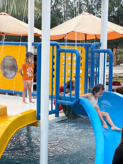 The scene depicts a water play area within a facility in Juwana, Indonesia. Three children are visible engaging with brightly colored water park equipment under daylight conditions. A young girl with dark, wet hair, wearing an orange short-sleeved shirt and light pink bottoms, stands barefoot on a wet, light-colored platform on the left. Water streams down around her from overhead fixtures. In the center, another child, mostly obscured by blue railing, wears red clothing and appears to be interacting with the water from within the play structure. To the right, a third young girl with wet, dark hair and a white tank top is positioned on a blue water slide, preparing to descend into the pool. A yellow slide is partially visible in the foreground on the left. The water park structure features yellow panels with circular cutouts and blue railings. White vertical poles support the overhead elements, from which water continuously falls. In the background, large, open beige and orange umbrellas provide shade, with dense green trees and a wooden deck visible beyond them. The pool water is disturbed by the play, showing ripples and splashes.