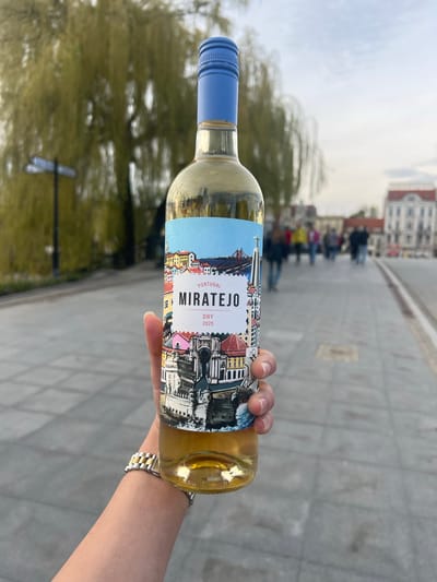 The image shows a hand holding a bottle of "Miratejo" white wine, a dry wine from Portugal, with its label featuring an illustration of city architecture. The person is wearing a silver and gold wristwatch. In the background, out of focus, there is a city view, including people walking on a pedestrian area with grey brick-like pavement, trees, and a building with white facade and red roof. Based on visual cues and common landmarks, it is most likely that the location is in the city of Bydgoszcz, Poland.
