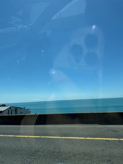 The image depicts a view from a vehicle, likely looking out through the windshield, overlooking the sea. The sky is a clear blue, with a few wispy clouds on the horizon. The sea is a vibrant turquoise color, stretching out to meet the sky. In the foreground, there is a road with a yellow line marking the lane. A dark concrete barrier separates the road from a building with a white facade and a red roof. The building appears to be situated on a slight elevation. Reflections are visible on the windshield, obscuring parts of the scene and showing what appears to be the interior of a car and potentially another vehicle outside. Based on the context of Khostinskiy, Russia, this scene likely captures a coastal view along the Black Sea, a common feature of the Khostinskiy district in Sochi.
