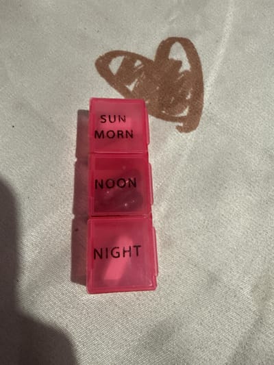 The image shows a pink pill organizer with three compartments labeled "SUN MORN," "NOON," and "NIGHT." The organizer is placed on a textured white surface, possibly fabric, which has a faint brown heart-shaped drawing on it. The lighting suggests an indoor setting, with a shadow cast on the lower left of the image. There is no indication of the location being in the city of Manila, Philippines.