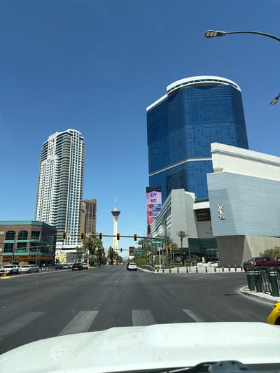 This image shows a street in Las Vegas, Nevada, USA. Prominent buildings include the Fontainebleau Las Vegas, the Wynn Las Vegas, and the Stratosphere tower in the distance. The view is captured from within a vehicle moving on the road. Traffic lights are visible. The weather appears sunny with a clear blue sky.