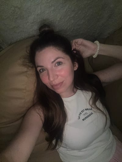 The image shows a young woman with dark hair pulled up in a bun, lying down and looking at the camera. She is wearing a white t-shirt with text that reads "LA VIE EST BELLE 1995 BORDEAUX". Her right arm is raised, and her hand is resting on her hair. Her left arm is bent at the elbow, with her hand resting on her chest. She appears to be indoors, possibly on a sofa or bed, with a textured surface behind her. The lighting is dim. The location is not specified in the image, and therefore cannot be determined to be Korsuntsi, Ukraine or any other city.
