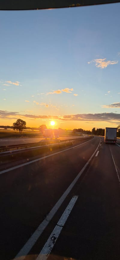 The image depicts a multi-lane highway near Bernau am Chiemsee, Germany, during sunset. The low-angle sun, positioned centrally on the horizon, casts an intense golden and orange glow across the sky and landscape, creating significant lens flare. The upper sky is clear blue with scattered, wispy clouds reflecting the sun's light.

On the left side of the highway, an open field appears covered in a thin layer of mist or fog, with a large, dark-leafed deciduous tree silhouetted against the bright horizon. The highway itself features at least two lanes in the direction of travel and a shoulder, delineated by distinct white markings, some showing wear. A metal guardrail runs along the median.

In the rightmost lane, a white semi-trailer truck is visible from the rear, traveling away from the viewer. Further ahead in the adjacent lane, a heavily sun-flared and partially obscured vehicle, likely a truck or large van, is also in motion. Additional distant vehicle lights are visible on the curving road ahead. The perspective suggests the image was captured from within a moving vehicle, looking forward through the windshield.
