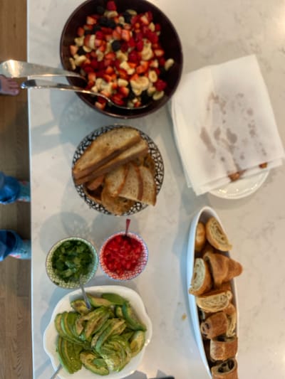 The image shows a countertop with a variety of food items arranged on it. The countertop is a light white marble with some light brown marble mixed in. There are two pairs of feet on the left of the image. The countertop is partially obscured by the feet. On the right side of the image there is a white paper towel with folds and some brown liquid stains on it. The food items are arranged on dishes and bowls in the center of the image. There is a bowl with a red rim that contains a fruit salad. The fruit salad contains chopped strawberries, bananas, blueberries, and raspberries. There is another bowl that contains a handful of sliced avocado. To the left of the avocado bowl there is a small bowl with sliced red tomatoes and another bowl with chopped cilantro. There is a white bowl with a black rim that contains a variety of croissants. There is a bowl with a black rim that contains a handful of sliced bread. The photo was taken inside an apartment or house. The location of the image is Rye, New York based on the latitude and longitude provided.