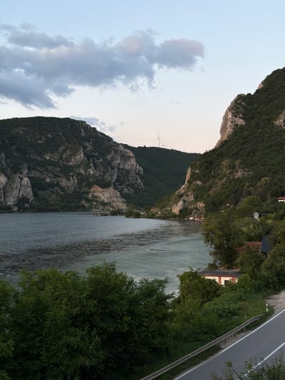 The image depicts a landscape view of the Danube River near the town of Golubac, Serbia. In the foreground, a paved road with a metal guardrail runs along the right side, bordered by dense green foliage. The road slopes downwards towards the river. The river itself occupies the middle ground, appearing wide and somewhat choppy, with what seem to be aquaculture nets or fish farms creating a textured pattern on its surface. Along the riverbank, several small buildings with red roofs and a warehouse-like structure with a white and orange facade are visible, nestled amongst lush green trees. In the background, imposing, rocky, and densely forested mountains rise steeply on either side of the river, creating a dramatic gorge. The sky above is a pale blue, with a few scattered clouds, and a distant wind turbine can be seen atop one of the hills. The overall impression is a natural, serene landscape with hints of human habitation along the river.