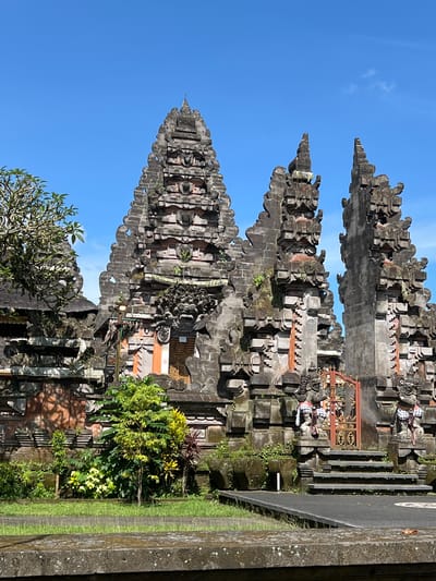 The image shows a Balinese temple in Petulu, Indonesia. Three tall, tiered, stone structures with intricate carvings are visible, set against a clear blue sky. The foreground features lush green vegetation and what appears to be a road with a low concrete barrier. Small figures adorn the temple structures, some wearing checkered cloths. The overall impression is of a well-maintained religious site in a tropical setting.