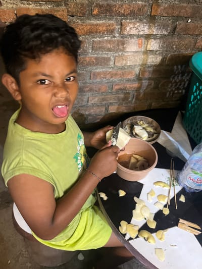 In the city of Mendoyo, Indonesia, a young boy with dark, curly hair is shown in a close-up shot, sticking his tongue out playfully at the camera. He is wearing a bright lime green t-shirt with a graphic on it, and a colorful bracelet adorns his wrist. The boy is seated and appears to be in the process of peeling a piece of fruit with a small knife. In front of him, on a black and white patterned table, are two bowls containing pieces of the peeled fruit, along with some wooden skewers. A bottle of water is also visible on the table, as is a green plastic basket in the background. The scene is set against a rustic brick wall, suggesting an outdoor or semi-outdoor setting. The lighting is somewhat dim, casting shadows and highlighting the textures of the brick and the fruit.