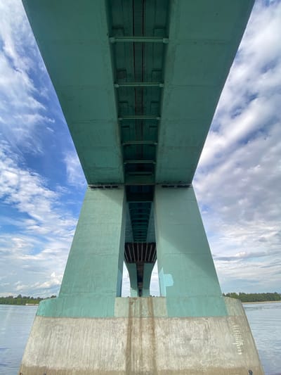 The image presents a striking upward-looking view of a large bridge structure, likely situated over a body of water in Grigor'evskoe, Russia. The dominant subject is the bridge itself, primarily painted in a faded teal or greenish-blue color. Massive concrete or painted metal beams create a complex interplay of horizontal and diagonal lines, forming a large "X" shape at a central junction point. The underside of the bridge reveals further structural elements, including visible crossbeams, supports, and railings.

On the far right side of the frame, a large, unpainted concrete or stone support pillar or abutment of the bridge stands out, its natural grey texture contrasting with the painted sections. Beyond the bridge structure to the right, a wide expanse of water is visible, likely a river or large lake, extending to a distant treelined shore. The upper and left portions of the image are filled with a bright, partly cloudy sky featuring wispy white clouds.

No human activity or specific event is taking place; the scene is static, focusing on the architectural grandeur of the bridge. The time of day appears to be bright daytime, with clear visibility. On the unpainted concrete abutment on the right, faint white markings are visible, appearing as a series of vertical lines and numbers, possibly "||||| |||||||11" or similar construction or survey indicators. The composition emphasizes the scale and robust engineering of the bridge against the backdrop of the sky and water.