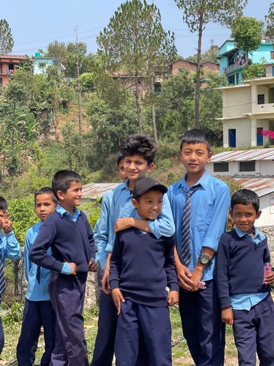A group of approximately six schoolboys in matching uniforms are closely grouped together, posing for a photo outside, in what appears to be Budar, Nepal. The boys are wearing light blue shirts, dark blue sweaters, and dark blue pants. One boy is wearing a black cap. Some are holding small, wrapped items in their hands. They are standing in a grassy area. The background contains residential buildings, trees, and other vegetation. The sky is clear and the lighting suggests it is daytime.