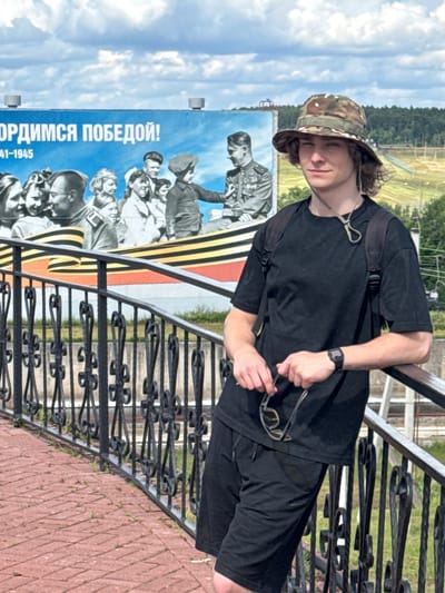 A young man with wavy, light brown hair and a camouflage bucket hat stands in front of a large billboard with a mural. He is wearing a black t-shirt and black shorts, and a black backpack is visible on his shoulders. He is leaning on a black metal railing and holding a pair of sunglasses. The billboard depicts a crowd of people, some in uniform, with the Russian text "ГОРДИМСЯ ПОБЕДОЙ!" ("We are proud of victory!") and the years "1941-1945" printed on it. The background shows a hilly landscape with trees and some structures, including a ski lift, suggesting a location outside of a dense urban area. This scene is located in Desnogorsk, Russia, at a memorial dedicated to Victory Day, likely near the Smolensk Nuclear Power Plant. The sky is cloudy.