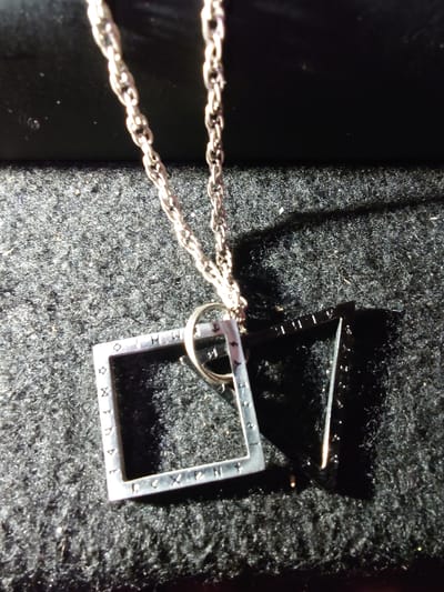 A silver chain with a square pendant is displayed on a dark, textured surface, possibly fabric. The pendant has a hollow interior and appears to have symbols or markings etched along its edges. A second, triangular frame is positioned partially behind and to the right of the square pendant, casting a shadow. The lighting in the image creates strong contrasts and highlights, suggesting an artificial light source. The scene is static and shows no signs of human presence or activity. The location in Sokolohirsk, Ukraine, is not visually indicated in the image.