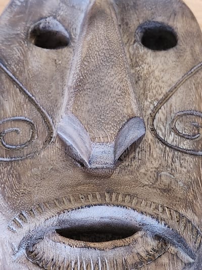 This is a close-up, overhead shot of a carved wooden mask. The mask is weathered and shows signs of age, with a textured, grainy surface. The carved details are intricate, featuring swirling patterns on the forehead and cheeks. There are two carved eye sockets, one of which is clearly visible, revealing a dark, deep opening. The nose is prominent and pointed, with a carved nostril on the visible side. The mask has a mouth that is not fully visible in this crop, but the carving suggests a closed or pursed expression. The overall color of the mask is a muted brown with some lighter, almost white, highlights where the carving is deepest or the wood has been worn. The texture of the wood is very apparent, with visible grain lines and a rough, unfinished feel. The mask appears to be an artifact, possibly decorative or ceremonial. There is no visible text in the image.