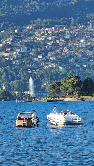 The image captures a vibrant, sunny day on Lake Como, Italy. The scene is dominated by the calm, blue waters of the lake in the foreground and midground, and a densely populated, green hillside in the background.
In the foreground, two boats are visible on the water. To the right, a white motorboat with a dark hull is present, with a person in a white shirt standing on the bow, looking towards the left. To the left, a smaller, dark-colored boat with wooden accents is seen, also with a person onboard. The water's surface is lightly rippled, reflecting the bright sunlight.
In the midground, a powerful water fountain sends a tall plume of white water high into the air, standing out against the backdrop of the city. The shoreline behind the fountain appears to have a promenade lined with trees.
The background showcases a sprawling residential area built into a steep, lush green hillside. Numerous buildings of varying sizes and colors, predominantly light-colored with visible rooftops, are nestled amidst abundant trees and foliage, extending far up the mountain. The entire scene is bathed in bright daylight, indicating clear and pleasant weather. No visible text is present in the image.