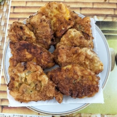 A white plate, lined with a white paper napkin, holds a serving of golden-brown, breaded cutlets. The cutlets appear to be chicken or fish, with a slightly irregular, crispy coating. They are arranged in a layered fashion on the plate, with some overlapping. The plate itself has a thin, dark line near its rim, bordered by a gold-colored band.
The plate rests on a placemat with a textured, wood-like pattern in shades of brown and beige. Behind the plate, the background shows a blurred image of a table setting with what appears to be a woven or bamboo-style placemat. The overall impression is that of a home-cooked meal. There is no visible text in the image. The lighting suggests it is daytime.