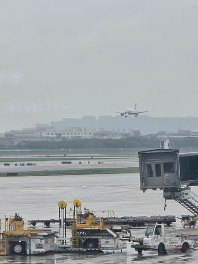 This image depicts an airport scene in Xiaoshan District, China, under a hazy sky. In the foreground, two yellow and white airport service vehicles are parked on the wet tarmac. One vehicle, on the right, is a white tow tractor with red accents, and another, on the left, is a larger white and yellow vehicle with a raised platform and wheels. A jet bridge extends from a building on the right side of the image towards the runway. In the middle ground, the wet tarmac of the airport runway is visible with various markings and some scattered black rectangular objects. In the background, a city skyline with low-rise buildings and trees can be seen through the atmospheric haze. An airplane, with its landing gear down, is descending towards the runway in the upper part of the image. The overall atmosphere is overcast, with wet surfaces suggesting recent rain. There are no people clearly visible in the image.