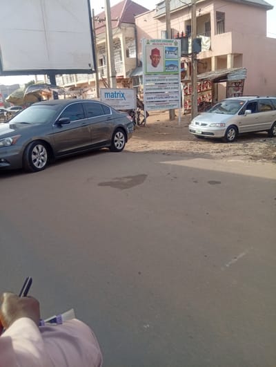 The image captures a street scene in Kaura Goje, Nigeria, showcasing two parked vehicles. A dark grey sedan is situated on the left side of the road, while a silver minivan is parked on the right. In the background, a building with signage for "CHOICE FASHION" and "SUPERMODEL DRESS" is visible. Further in the background to the right, is a pinkish building.
A sign displaying "KANO STATE GOVERNMENT" and "KANO STATE MINISTRY OF EDUCATION" can be seen near the silver minivan. A smaller sign reading "matrix Gas" is positioned near the dark grey sedan.
A person can be seen in the foreground, holding an object that appears to be a pen. The environment seems dusty and somewhat cluttered.