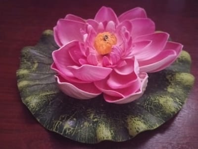A close-up, overhead shot shows a faux pink lotus flower resting on a dark green, textured lily pad. The flower's petals are a vibrant pink, with a yellow stamen at its center. The lily pad is wavy and has a mottled green and yellow appearance. The objects are placed on a dark brown, wooden surface, possibly a table. The lighting is dim, casting soft shadows and highlighting the textures of the objects. The image does not contain any information or visual cues that would indicate a location in Lingiai, Lithuania, or any specific urban environment.