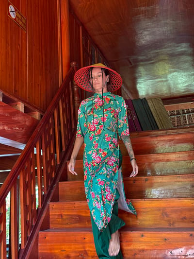 An individual is captured in a posed photograph within a warm, wooden interior, likely a traditional house or building in Thông Nguyên Commune, Vietnam. The person is positioned lying down or leaning across a set of wooden stairs, looking directly at the camera with a calm, neutral expression.

The subject is wearing a vibrant green garment, possibly a dress or tunic, adorned with a floral pattern of red, pink, and white flowers with green leaves. Underneath, green pants are visible. A distinctive red, woven straw hat with a yellow rim covers their head. The person has dark hair, styled in braids or dreadlocks, visible beneath the hat. A full sleeve tattoo is prominent on their right arm. Their left hand, showing manicured nails, is extended towards the left side of the frame.

The setting is characterized by extensive use of polished wooden planks for walls and a robust wooden staircase with intricately carved or slotted railings and steps. In the upper left background, stacked items resembling folded textiles or mattresses in various muted colors (olive green, dark green, burgundy) can be seen against a lighter, textured wall. The lighting suggests daytime, with natural light reflecting off the wooden surfaces, creating bright patches.

A "NO SMOKING" sign is affixed to the wooden wall in the lower left foreground. The sign features a red circle with a diagonal line through a cigarette symbol and includes text in both Vietnamese ("CẤM HÚT THUỐC") and English ("NO SMOKING").