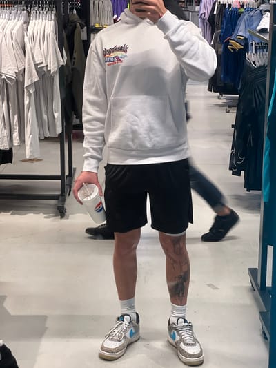 A man stands in what appears to be a clothing store in Swindon, United Kingdom, holding a disposable cup with a straw. He wears a white hoodie with a logo, black shorts, white socks, and patterned sneakers. His legs are tattooed. Another person is visible in the background, walking. Clothing racks with folded and hanging garments are visible on the left side. The floor is a light gray or white.