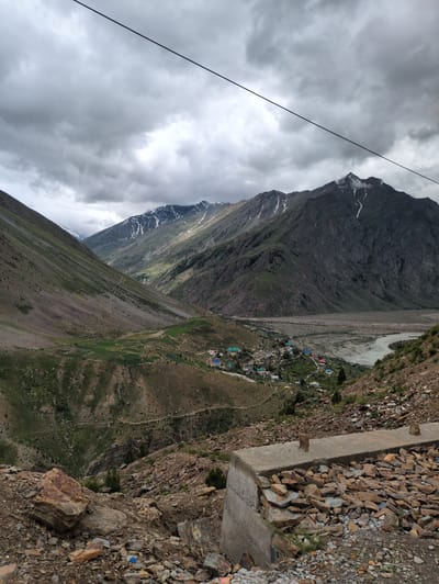 The image shows a village nestled in a valley in Darcha, India, surrounded by tall, rugged mountains. The sky is overcast with dramatic, dark clouds. A power line stretches diagonally across the upper part of the frame. The foreground is dominated by a concrete structure filled with stones, typical of infrastructure in such a mountainous region. The village itself appears to be a collection of small, colorful houses with some agricultural fields visible on the slopes. A river flows through the valley in the middle ground. The overall scene depicts a remote, high-altitude settlement in a dramatic natural landscape.