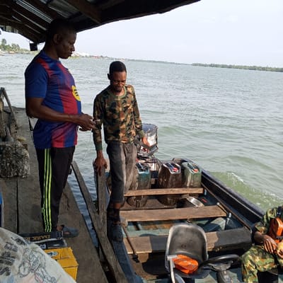 The image depicts an outdoor scene at a wooden dock in Koko, Nigeria, adjacent to a wide body of water with a clear sky overhead and a distant forested shoreline. A wooden overhead structure partially shades the dock area.
Three adult males are present. On the left, a man wearing a blue and red striped short-sleeve shirt, black track pants with green stripes, and dark slip-on shoes stands on the wooden dock, holding a white rectangular object in his hands. To his left, a partially visible white sack marked 'DARK R' and a yellow box are on the dock.
In the center, a second man wearing a camouflage long-sleeve shirt and dark jeans is positioned with one foot on the dock and the other inside a dark utility boat. He leans on the boat's gunwale with his left hand. The boat, equipped with a silver and black outboard motor at its stern, contains at least three green plastic jerry cans, a metallic bowl, and a dark plastic seat with an orange object on it.
A third man, partially visible on the far right and seated in the boat, is wearing a camouflage shirt and holding a light-colored object in his hands. The water exhibits minor surface ripples. The scene illustrates maritime activity, likely involving transport or refueling.