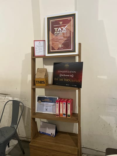 The image depicts a shelf display in what appears to be a cafe or business establishment in Makassar, Indonesia. The shelf is made of light-colored wood and is filled with various items.

On the top shelf, a framed certificate or award is prominently displayed, featuring Indonesian text and the title "TAX AWARD 2025". Below it, to the left, is a smaller framed document.

The second shelf holds two cardboard boxes, one stacked on top of the other. The top box is brown and has text that includes "POTENTIAL OPTIMIZATION VALUE" and "POV". The bottom box is yellow and has "THIS IS" written on it in large, bold letters. To the right of the boxes is a dark-colored rectangular sign with "DELIFRU DRINK EXPERT" at the top and "CONGRATULATION 2nd Runner Up IDR 500 THOUSAND" in the center.

The third shelf displays a framed certificate or award with Indonesian text that reads "PIAGAM PENGHARGAAN" and mentions "KOPITEORI MANDIRI INDONESIA". To the right of this certificate are four identically shaped and sized boxes, vertically stacked. They appear to be product packaging and have visible text in red and white.

The bottom shelf holds a desk calendar with Indonesian text and an image of a family or group of people.

The background of the scene is a light-colored wall, showing some texture and possible imperfections. To the left of the shelves, a dark gray metal chair is visible. The overall impression is one of an organized display of achievements and possibly products.