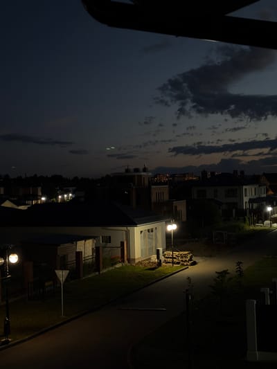 The image depicts a twilight scene in the village of Martemyanovo, Russia. The sky is dark with scattered clouds, and artificial lights illuminate the environment. In the foreground, a street lamp with a white globe is visible, casting light on a paved road and surrounding grassy areas. To the left, a building with white walls and glass windows is partially visible. Further into the scene, several houses with illuminated windows and exterior lights are scattered throughout the landscape. In the distance, a cluster of taller buildings with glowing lights suggests a more urban area. The overall impression is of a quiet residential neighborhood at dusk or early evening.