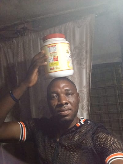 A man balances a large, off-white container with a red lid on his head, holding it in place with his left hand. He wears a black mesh shirt with orange and white stripes on the sleeves and collar. He has short hair, dark skin, and several scars on his face. Background elements include a curtain and what appears to be an air conditioning unit. The lighting is dim.