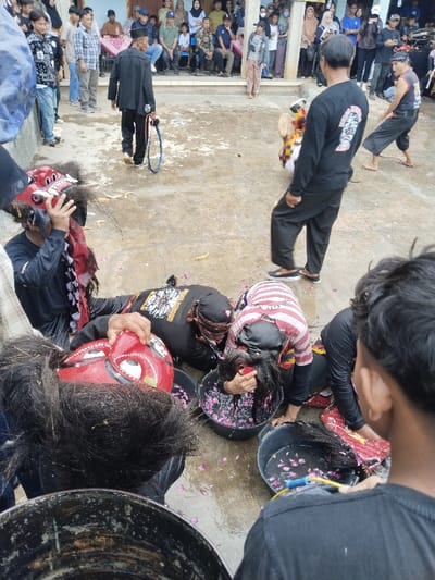 This image depicts an outdoor cultural performance or ritual in Kerjo, Indonesia. In the foreground and mid-ground, multiple individuals are actively engaged. Three male participants are prominently bent over, submerging their heads or dark hair into black plastic buckets containing water and scattered pink flower petals. One individual holds a large, red, stylized mask with white eyes and black hair near their face, while another identical mask rests on the ground beside a bucket.
Other participants include a male in a black long-sleeved shirt and black pants standing upright, observed from the back. To his right, another male in a dark sleeveless top and loose pants is in a crouched or active stance. In the background, a male wearing a black jacket and pants walks away from the central activity, carrying a blue braided whip or rope. A performer in a yellow and white costume, possibly an animal mask, is visible in motion further back on the right.
Approximately 30-40 spectators, including men, women, and children, are visible in the background, seated and standing on a raised concrete platform or porch of a light-colored building, observing the event. The ground is a wet, grey concrete surface with scattered pink flower petals and some larger yellow organic debris. The scene is illuminated by natural daylight.