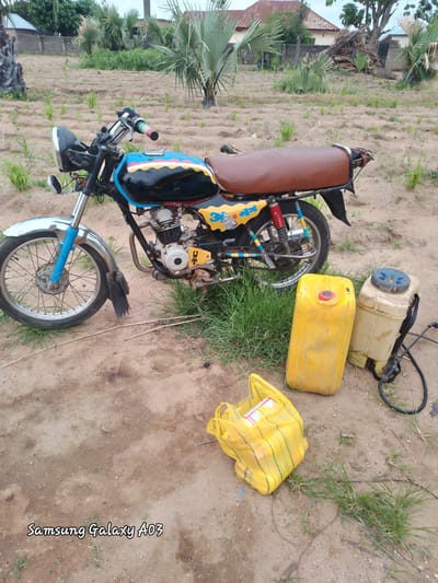 The image depicts an outdoor scene in Angwan Hausawa, Nigeria. A motorcycle, painted with black, blue, yellow, and red stripes on its fuel tank, is parked on a dirt ground. The motorcycle has a brown seat and a chrome front fender. To the right of the motorcycle are two yellow plastic containers and a yellow plastic bag. One of the containers is tall and cylindrical with a red cap, while the other is shorter and rectangular with a blue lid. The yellow plastic bag, filled with items, is positioned in the foreground. The environment consists of dry, sandy soil with patches of green grass. In the background, palm trees and some buildings with red roofs are visible. A watermark at the bottom left corner reads "Samsung Galaxy A03".