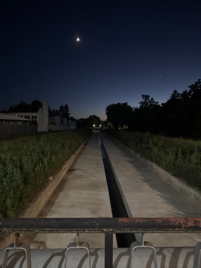 The image captures a long, concrete canal at night in Popovo, Bulgaria. The canal is flanked by overgrown grassy embankments and trees on either side. In the distance, lights from buildings and street lamps illuminate the scene. The sky above is dark, with a crescent moon and a faint star visible. The foreground shows a metal railing, suggesting the photo was taken from a bridge or elevated vantage point. The overall atmosphere is serene and quiet.