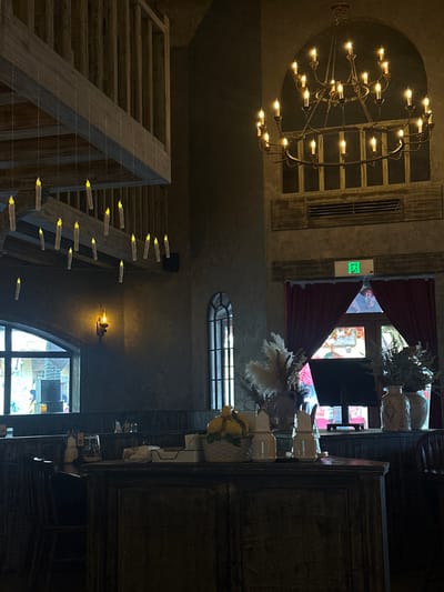 The image depicts the interior of a dimly lit, themed establishment, likely a restaurant or cafe, located in Licheng District, China. The scene has a gothic or fantasy aesthetic.

A prominent feature is a large, two-tiered metal chandelier with approximately two dozen illuminated faux candles, suspended from the ceiling in the upper right quadrant. Below it, an architectural recess and a horizontal wooden panel with a dark air vent are visible. In the upper left, a wooden balcony railing with vertical balusters extends across the frame. Below this railing, numerous faux "floating candles" with yellow flames are suspended at varying heights by thin, translucent lines.

The walls are textured, appearing dark gray or brown with a distressed finish. On the left, a wide arched window provides a view of a brighter exterior, possibly a street or courtyard, with faint colors and shapes. A single wall sconce with a warm, glowing bulb is mounted on the wall below this window. A tall, narrow arched window with vertical mullions is situated further right.

In the foreground, a dark, rustic wooden counter or cabinet runs horizontally. Upon this counter are various objects: several clear glasses, small condiment shakers/bottles, a tissue dispenser, a white woven basket containing yellow decorative items resembling lemons, and two white ceramic dispensers. Behind these items, two large vases hold dried floral arrangements, including light-colored pampas grass.

In the background, beyond the counter, red velvet-like curtains are partially drawn over a glass doorway or large window, revealing a brighter area beyond. Above these curtains, a green illuminated "EXIT" sign, including Chinese characters, is visible. Partial views of dark wooden chair backs are visible on the far left and right. No individuals are present in the observable frame.
