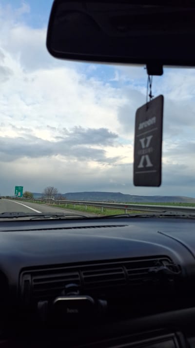 The image is taken from inside a car, looking out the front windshield. A rear-view mirror and an air freshener hang from the top of the windshield. The outside scene features a cloudy sky and a road with green vegetation and a distant mountain range on the right. A green road sign is visible on the left side of the road. The road appears to be a highway. Based on the road sign "Златина 29 км" (Zlatina 29 km), the car is located approximately 29 kilometers away from the city of Zlatina, Bulgaria. The car's dashboard and air vents are visible on the right side of the frame.