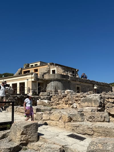 The image captures a sunny day at the Minoan palace complex of Knossos, located on the outskirts of Heraklion, Crete, Greece. The scene is dominated by the ancient stone ruins of the palace, bathed in bright sunlight. In the foreground, a woman wearing a straw hat, white t-shirt, and a purple skirt walks across a stone path, carrying a small brown purse and a green bottle. Behind her, to the left, two men are visible. One man, wearing a white t-shirt and shorts, stands facing forward, while another man in a black t-shirt is partially visible behind him. In the background, the reconstructed sections of the palace stand with their yellow and stone facades. Two individuals are standing on a higher level of the ruins, one appearing to take a photograph. The sky above is a clear, vibrant blue. The environment consists of weathered stone structures, pathways, and natural vegetation, indicative of an archaeological site.
