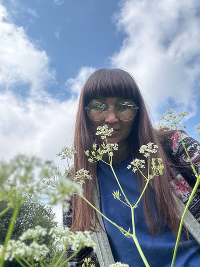 A low-angle, medium shot captures a woman with long, dark brown hair and bangs, wearing round, reflective sunglasses and a blue t-shirt with a patterned cardigan. She is surrounded by white wildflowers in the foreground and appears to be outdoors with a cloudy blue sky in the background. The perspective suggests she is looking directly at the camera. The scene is set in a natural environment, likely a field or meadow, in the village of Sredneye Kechevo, Russia.