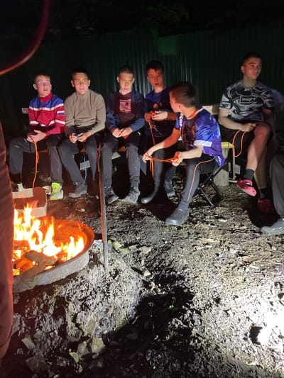 In Nikolaevka, Russia, six young males are seated outdoors at night around a bonfire. The bonfire is contained within a metal ring on the ground, which is covered in dark soil and small stones. The males are seated on a wooden bench and folding chairs, facing the fire. They are dressed in casual clothing, including t-shirts, jackets, and pants. Several of the males are holding orange ropes, and their expressions suggest they are engaged in an activity or conversation. The background is dark and appears to be a natural setting, possibly a backyard or campsite, with a dark fence or wall visible. The lighting is primarily from the bonfire, casting a warm glow on the scene and creating shadows.