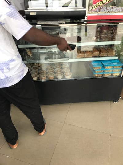 The scene is an interior view within the city of Calabar, Nigeria, featuring a person interacting with a food display case. A dark-skinned individual, observed from the waist down and partially from the back, is visible on the left. The person wears a white short-sleeved shirt with light blue horizontal stripes, black trousers, and light blue and orange open-toed slippers. Their right arm extends into a multi-tiered glass display case, with a black smartphone held in their right hand, positioned over a silver tray.
The display case, featuring a black base and frame, contains various food items. The top section, partially visible, includes a red and white sign displaying the word "POPCORN" and what appears to be popcorn. The middle shelf, accessible to the person, holds multiple individually wrapped rectangular loaves, possibly bread or cake, and other packaged goods. The bottom shelf displays numerous small, round clear plastic containers with white lids, arranged in rows, containing what appear to be individual desserts or snacks. Adjacent to these are stacks of clear rectangular plastic containers with blue lids, filled with fried plantain chips or similar items. The display case rests on wheels. The flooring consists of light-colored square tiles with dark grout lines.