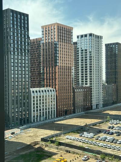 The image shows a high-angle view of several tall buildings in an urban environment. The buildings are characterized by repetitive window patterns and varying colors: dark gray, brown, and light gray. Adjacent to the buildings is an unpaved area with rows of parked vehicles, primarily white in color. Some construction or excavation activity is visible in the lot with a yellow vehicle nearby. The sky is visible in the background with scattered clouds. Based on the prompt, the location of this image is Ukromne, Ukraine.