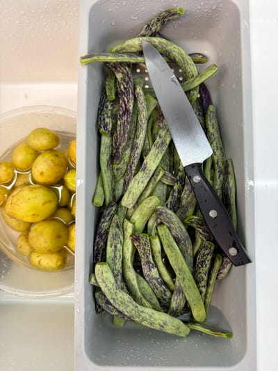 The image shows a gray container filled with dragon tongue beans, also known as borlotti beans. A knife rests on top of the beans. Below the container of beans is a clear plastic container with potatoes submerged in water. The image was captured in the city of Arinj, Armenia.
