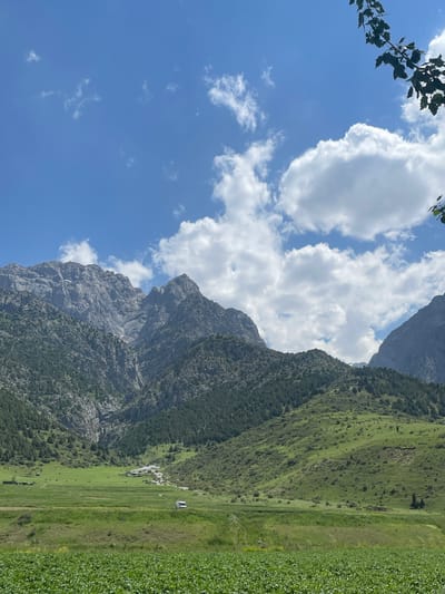 The image depicts a sweeping mountain landscape under a bright blue sky dotted with fluffy white clouds. In the foreground, a lush green field, possibly cultivated with crops, stretches across the bottom of the frame. Behind this, a vast expanse of rolling green hills, densely covered with dark green trees, rises to meet a dramatic, rugged mountain range. The mountains are characterized by sharp peaks, steep slopes, and rocky textures, with some lighter patches suggesting snow or scree. In the mid-ground, nestled in a valley between the hills and mountains, are a cluster of small buildings and a road or track. A white van is visible on the road. The overall impression is one of natural beauty and rural serenity in a mountainous region. This scene is likely located in or near the city of Khaidarkan, Kyrgyzstan, a region known for its mountainous terrain and natural landscapes.