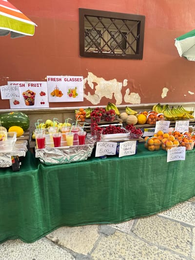 This image captures a vibrant outdoor fruit and juice stall set against a weathered reddish-orange stucco wall in Bari, Italy. The wall, showing numerous irregular patches of peeling paint that reveal lighter plaster underneath, features a dark brown-framed window with decorative metal security bars positioned above the center of the stall.
Two laminated white signs are taped to the wall above the fruit display. The left sign, titled "FRESH FRUIT," includes a handwritten note "ANGURIA 2,00 Kilo" (Watermelon 2.00/kg) and images of a fruit salad cup, strawberries, and cherries. The right sign, titled "FRESH GLASSES," displays images of fresh orange juice and a green juice. Partially visible at the top left is a segment of an orange, white, and lime green striped market umbrella, while a green and white striped umbrella is seen at the top right.
The market stall itself is draped with a dark green, finely meshed fabric tablecloth that extends close to the ground. On the left side of the stall, a large dark green watermelon and a bright yellow melon rest on a wooden crate, adjacent to a clear plastic container. The central area of the stall showcases a row of at least six clear plastic cups, each containing a brightly colored fresh juice or smoothie (red, light yellow, and orange) and a light blue straw. These cups are arranged on a base covered in crinkled aluminum foil, likely to keep them chilled. Behind the juices, several piles of green-yellow fruit, possibly pears or a different type of melon, are visible.
Moving to the right, the stall is laden with various fruits displayed in clear plastic containers or arranged on wooden crates. These include numerous clusters of ripe yellow bananas, bright red strawberries, apricots, cherries, and peaches or nectarines. Each fruit type has a handwritten price sign in Italian, indicating the price per kilo or per basket: "ALBICOCCO 4,00 Kilo" (Apricot 4.00/kg), "BANANE 2,50 Kilo" (Bananas 2.50/kg), "FRAGOLE 5,00 Cestino" (Strawberries 5.00/basket), "CHERRY Ferrovial 7,00 Kilo" (Cherries, Ferrovial variety 7.00/kg), and "PERICOCI SELEZIONATI 5,00 Kilo" (Selected Peaches/Nectarines 5.00/kg).
The ground in the foreground consists of uneven, light-colored stone paving slabs, characteristic of an old street or pedestrian area. No people are visible in the frame.