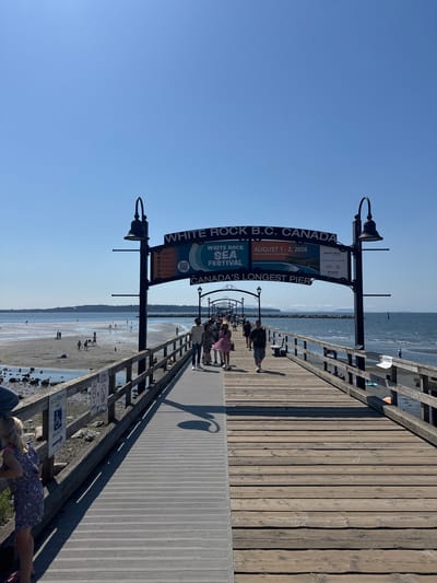 The image depicts a sunny day at White Rock's pier, recognized as Canada's longest pier. In the foreground, a young girl in a striped dress stands by a wooden railing. The pier itself is a broad walkway, divided into two sections: a grey metal pathway on the left and a weathered wooden deck on the right. People are strolling along the wooden portion of the pier, some in casual attire, others in more festive clothing.
To the left of the pier, a sandy beach is visible, dotted with various rocks and shallow water. Several people are scattered across the beach, some walking, others appearing to be sunbathing or playing. In the distance, across the water, a tree-lined shoreline can be seen, suggesting the presence of land beyond the immediate coastal area.
Above the pier, a prominent sign announces "WHITE ROCK B.C. CANADA" and promotes the "WHITE ROCK SEA FESTIVAL" scheduled for August 1-2, 2026, to be held at Memorial Park & West Beach. The sign also lists activities such as "Torchlight Parade," "Fireworks," "Car Show," "Kids' Activities," "Food Trucks," and "Live Music." Beneath this, it reiterates that this is "CANADA'S LONGEST PIER." Two ornate lampposts with bell-shaped shades stand on either side of the main sign, adding to the pier's classic aesthetic. The sky is a clear, vibrant blue, indicating fair weather conditions. The overall scene is one of a bustling public space enjoying a pleasant day by the sea.
