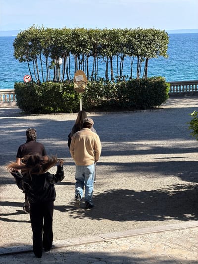 The image shows a scene along the Lungomare coastal promenade in Opatija, Croatia. The Adriatic Sea is visible on the left side of the image. A row of cultivated trees lines the promenade, with a "no swimming" sign affixed to one tree. Three individuals are walking on the paved path. The first person is wearing a light-colored corduroy jacket and jeans. The second person, in the middle, has long hair and is wearing a dark-colored top. The third person, closer to the viewer, is dressed in black. Shadows cast by the trees and the people are visible on the path. The perspective is from above and slightly to the side.