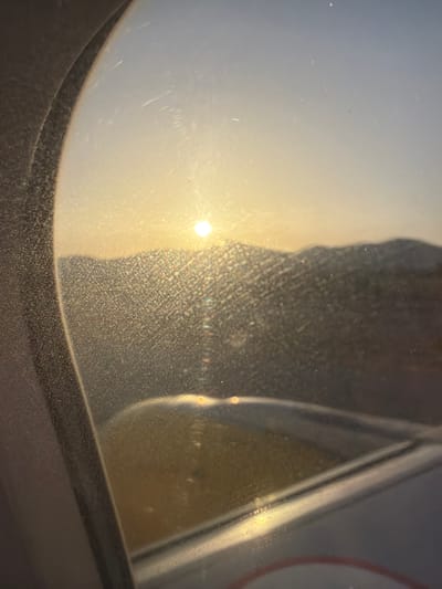 The image shows a view from an airplane window during sunset. The sun is visible in the upper center of the frame, casting a warm glow on the clouds and distant mountains. The windowpane is dirty and scratched, creating a hazy effect. A portion of the airplane wing is visible in the foreground. The scene is likely in the vicinity of Sant Josep de sa Talaia, Spain, given the mountain landscape often seen in the area during flights. There are no people or specific objects identifiable in the image beyond the airplane's interior and exterior elements, and the natural landscape. The overall impression is one of a serene, albeit slightly obscured, sunset viewed from a high altitude.