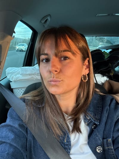 A female individual is depicted in the foreground, seated inside a vehicle, presumed to be in Bourg-lès-Valence, France. She has shoulder-length brown hair with bangs, light skin, and is looking directly at the camera with a puckered lip expression. She is wearing a white top, a blue denim jacket, and a gold-toned, textured hoop earring in her right ear. A dark seatbelt is secured across her chest, extending diagonally from her left shoulder to her right hip. In the left background, a white perforated laundry basket filled with white fabric is visible on an adjacent seat. To her right, in the rear of the vehicle, multiple dark boots are stacked. Through the vehicle's left side window, a multi-lane highway is visible, with a white sedan in the distance and a roadside barrier. The lighting conditions suggest either early morning or late evening.